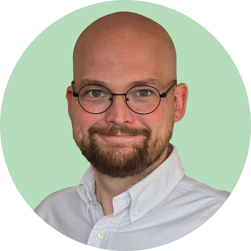 Smiling bald man with a beard and round glasses wearing a white button-up shirt against a light green background.