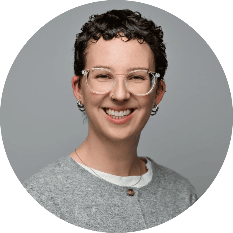 Smiling person with short curly hair, clear-framed glasses, hoop earrings, and a grey sweater over a white shirt.