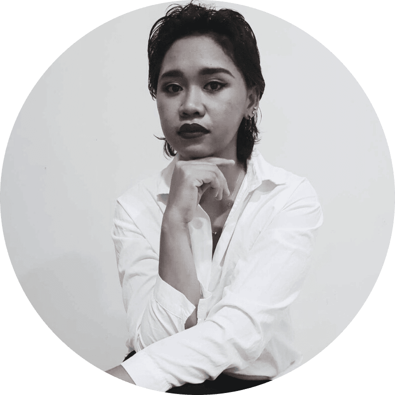 Person with short dark hair wearing a white shirt, resting their chin on a hand, looking thoughtfully at the camera.