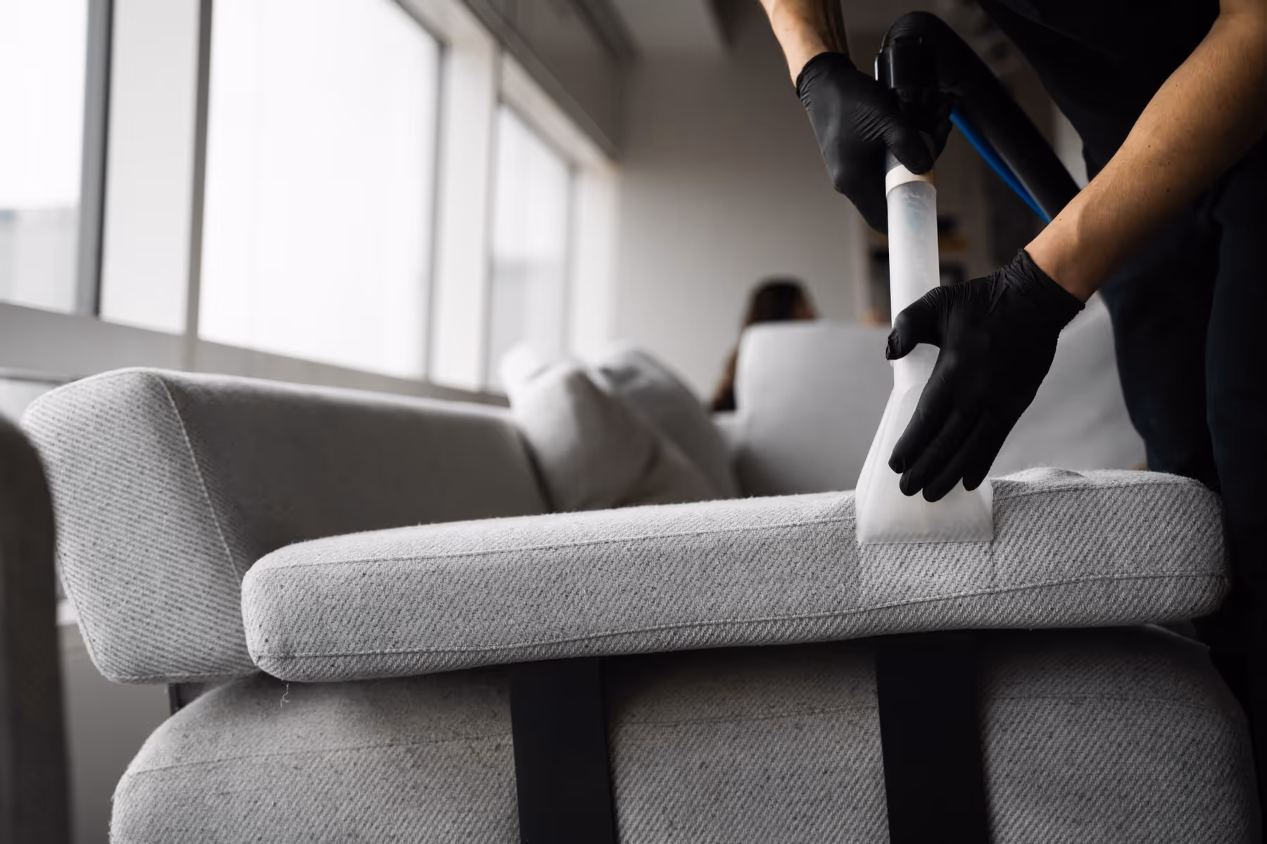Person wearing black gloves steam-cleaning a gray fabric sofa cushion near large windows.