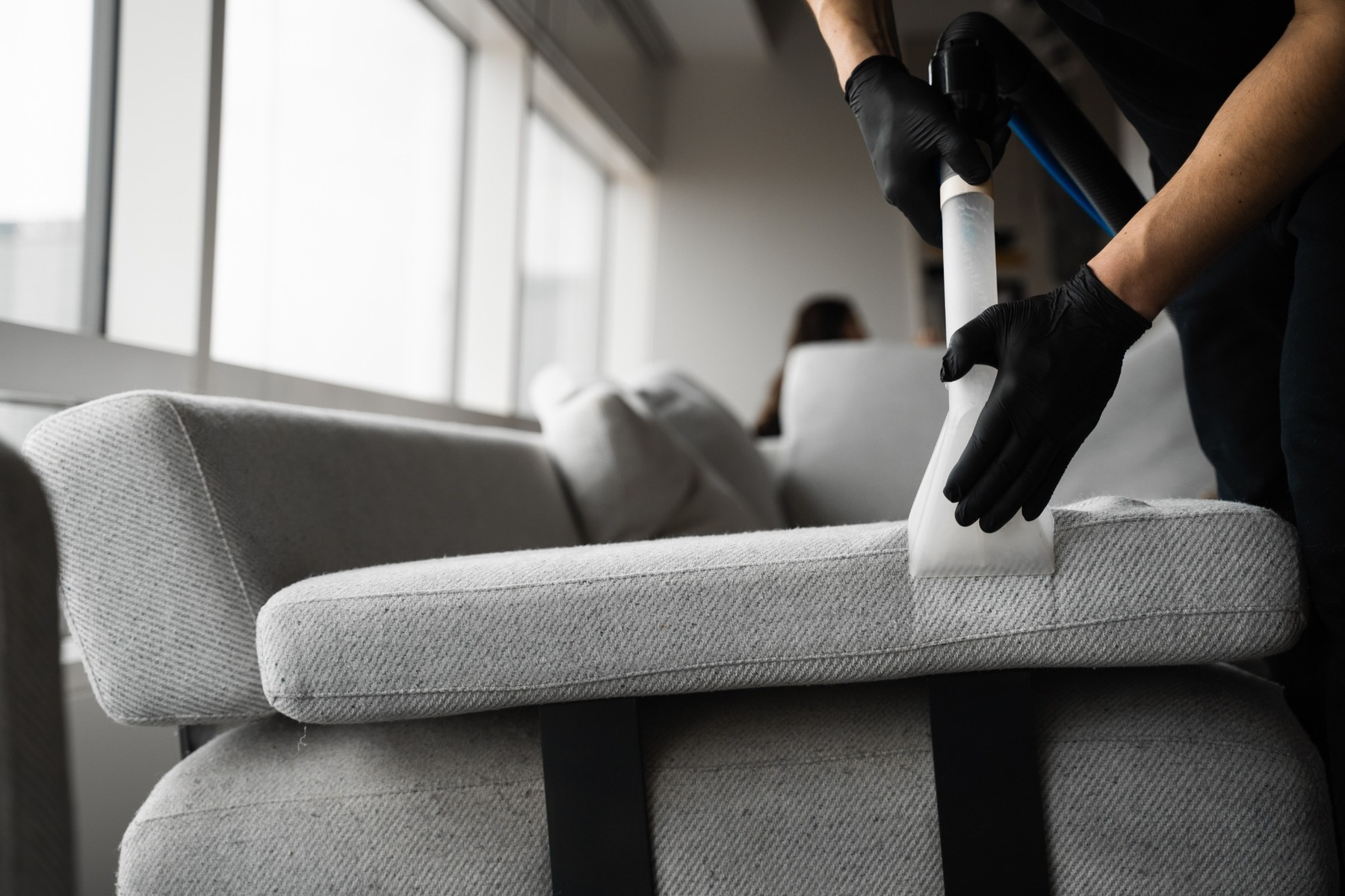 Person wearing black gloves steam-cleaning a gray fabric sofa cushion near large windows.