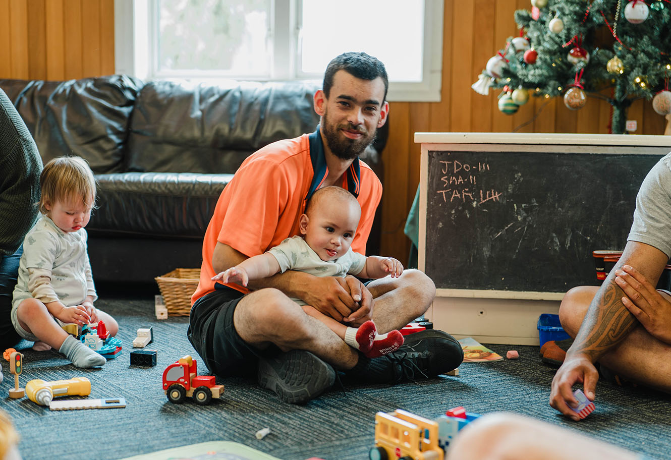 a father plays with a young child on the floor with other family nearby