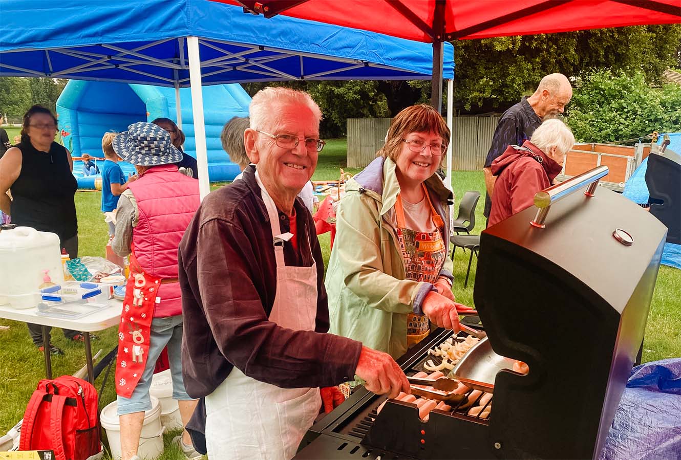 a man and woman serve sausages and other food from a barbecue under colourful gazebos