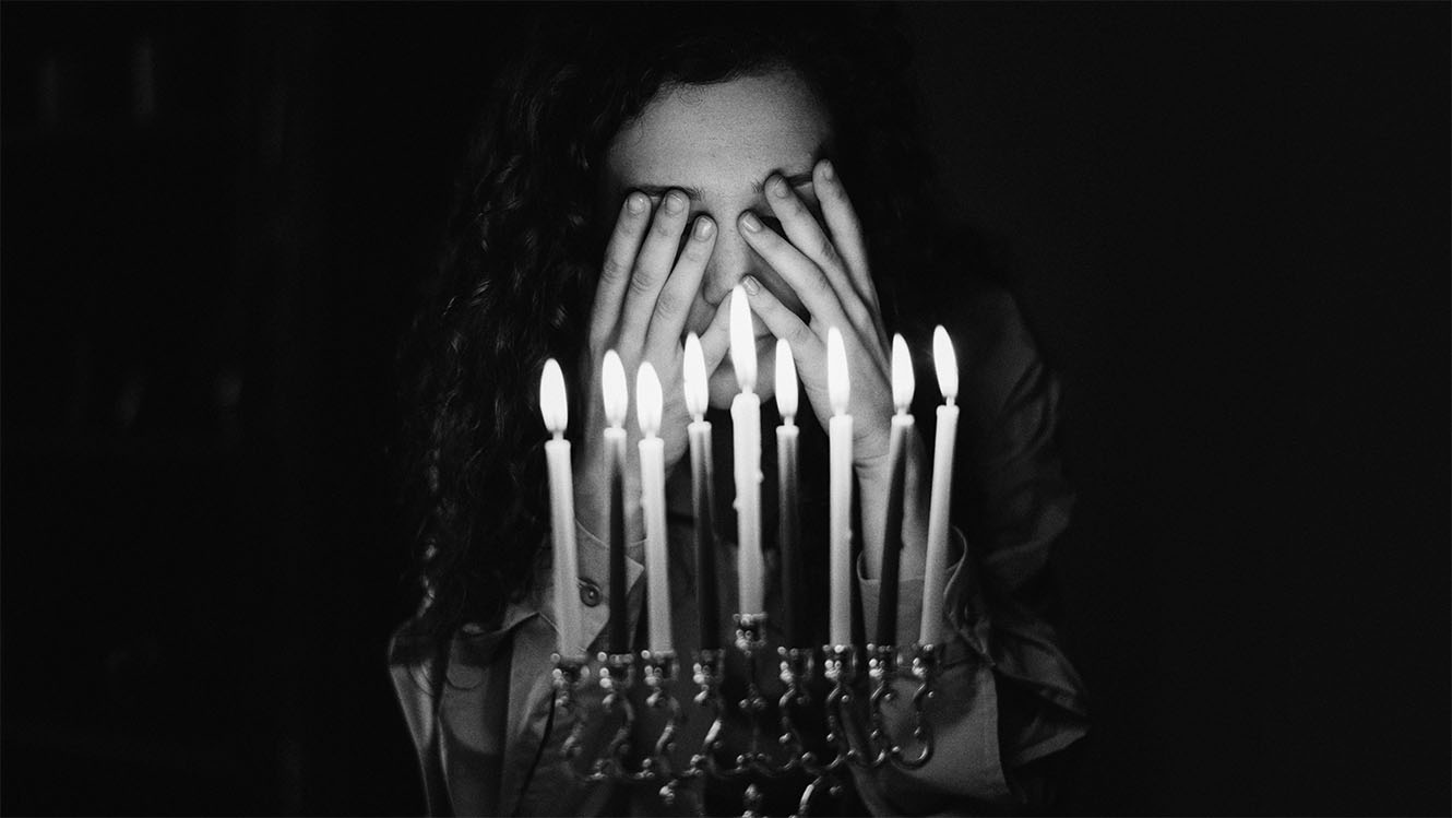 a person covers their eyes behind a lit menorah