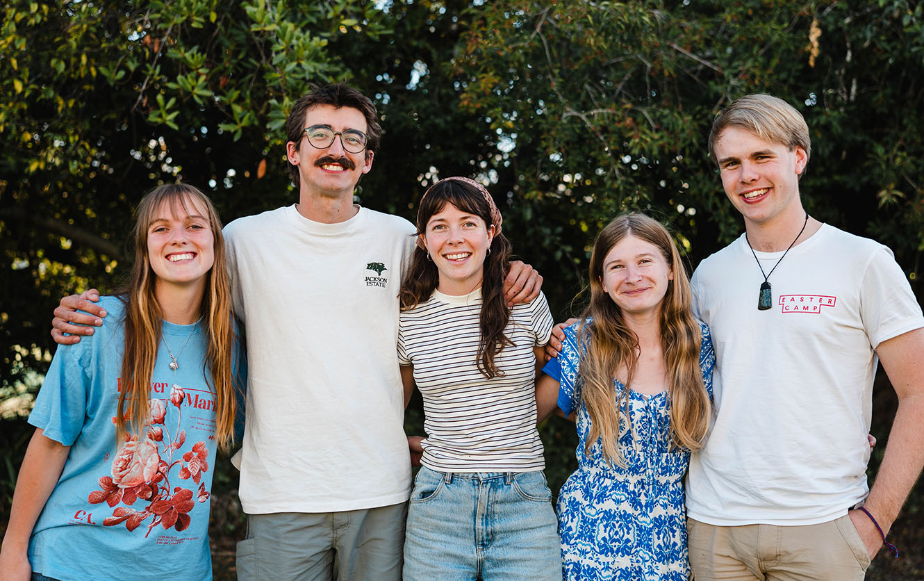 The members of the St Barnabas vicarage flat with their arms around each other, outside in front of some foliage.