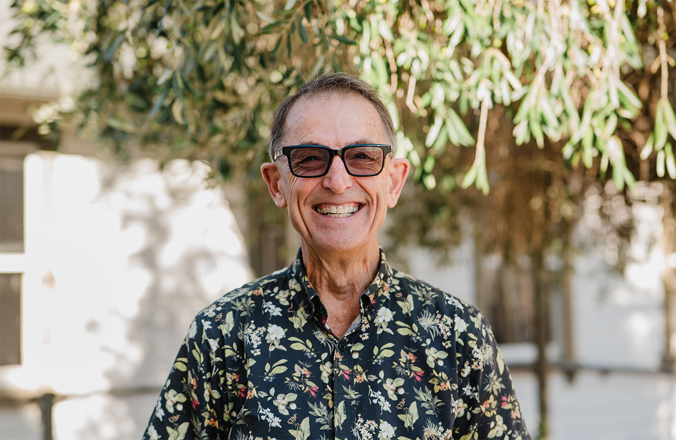Head and shoulders of David Allpress, smiling outside All Saints church in front of an olive tree.