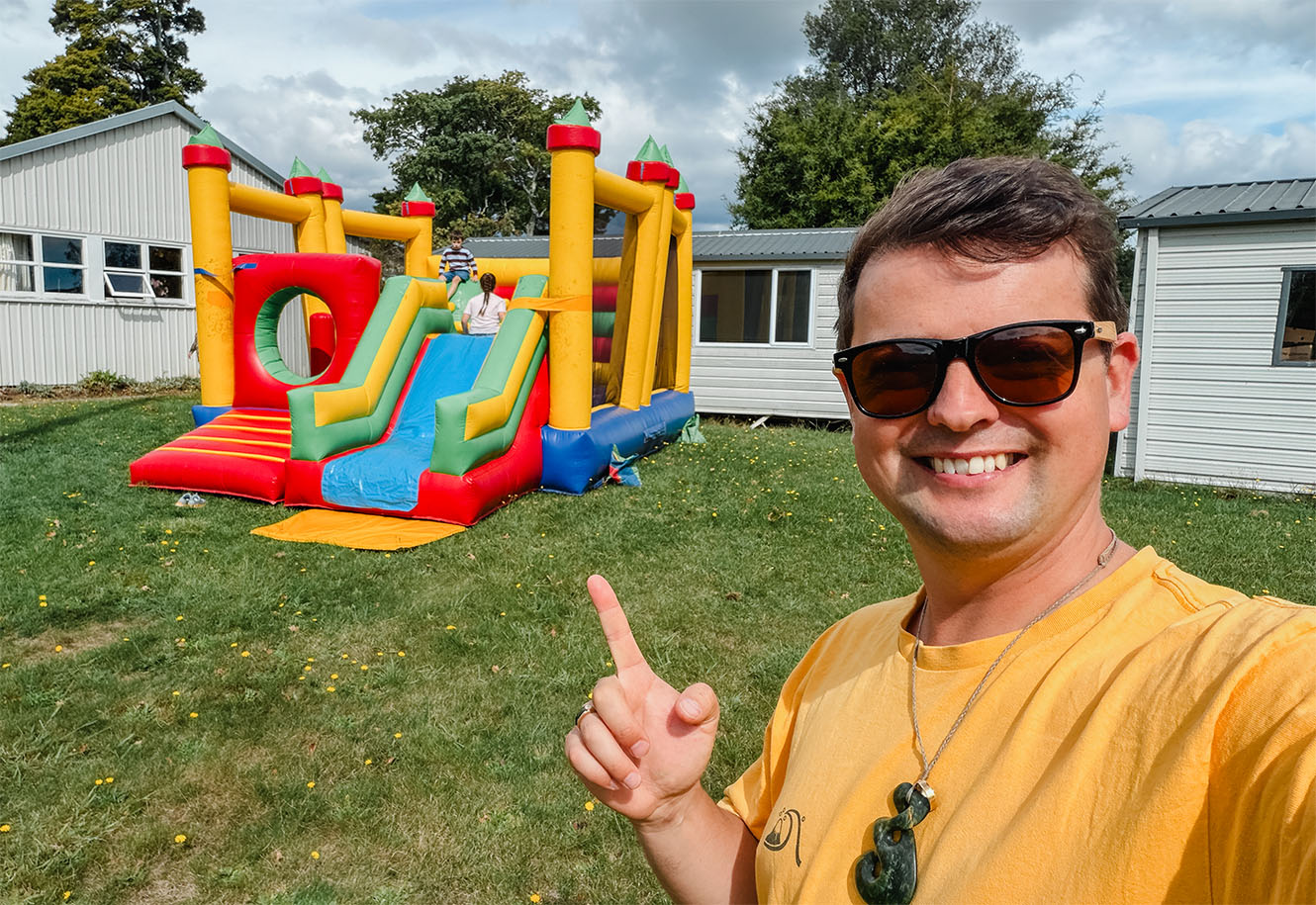 Rev Brad Wood smiling for a selfie in front of a bright bouncy castle next to St Paul's in Brightwater.
