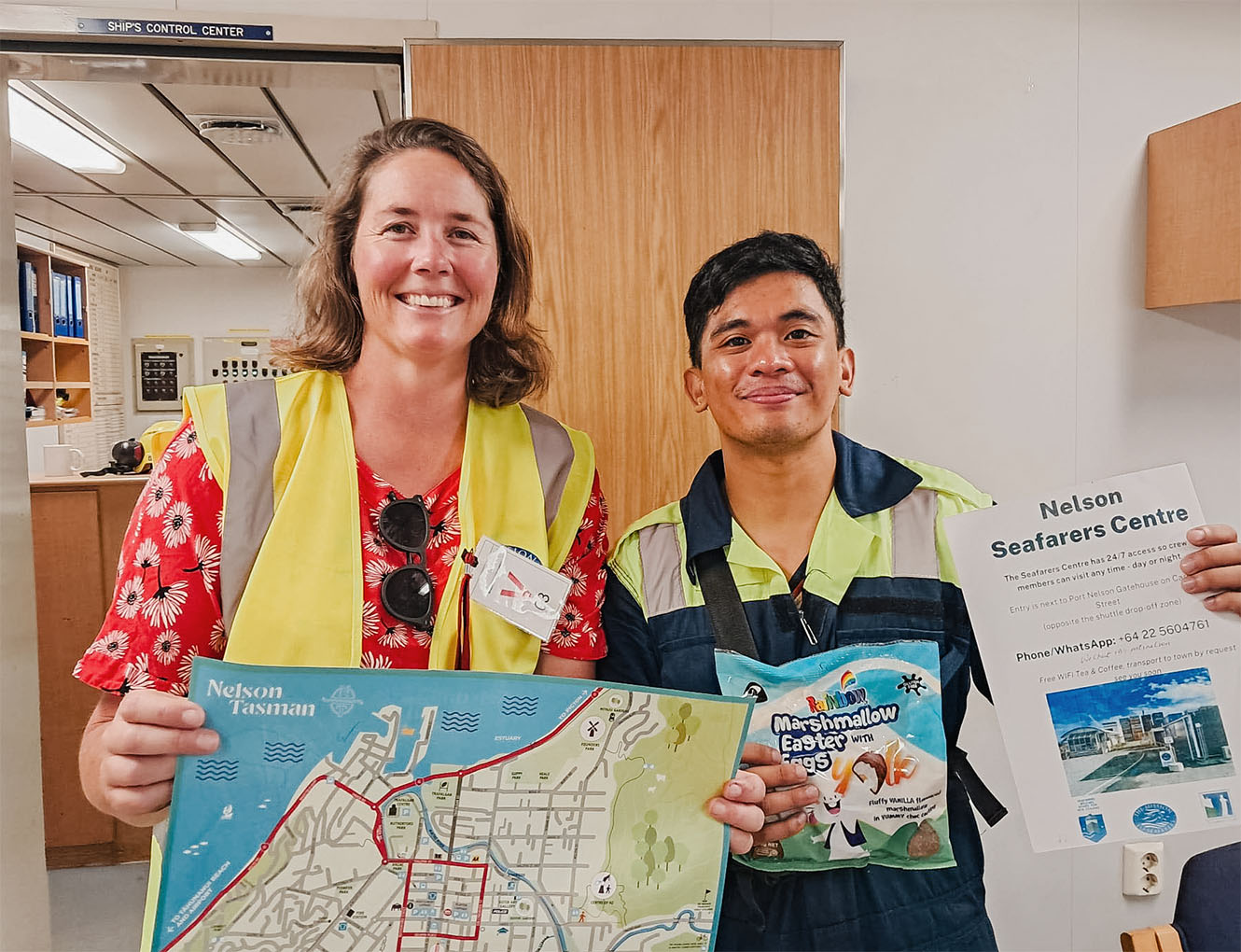 Amba holds a map of Nelson while a man adjacent presents an informational poster about the Nelson Seafarers Centre.