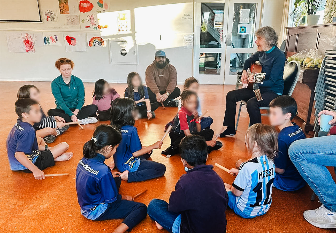 Maria plays guitar with a group of kids sitting on the floor around her, listening