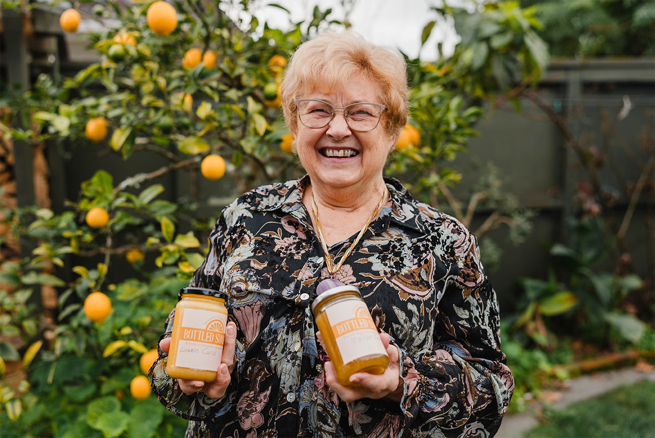 Sheena Pearson smiles in front of a lemon tree, holding two jars of preserves with the labels "Bottled Sunshine"