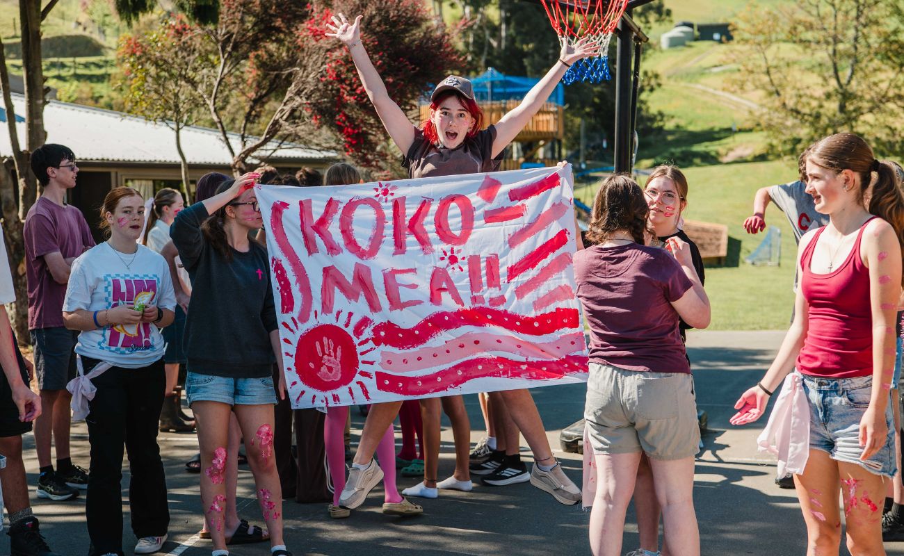 A girl jumps up behind a large white flag painted with pink, saying "Kokomea"