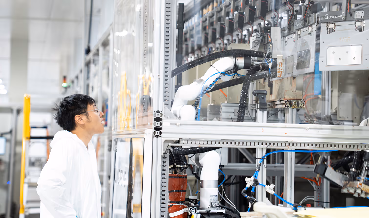 Technician in white coat closely inspecting complex robotic machinery with tubes and cables in a laboratory or factory setting.