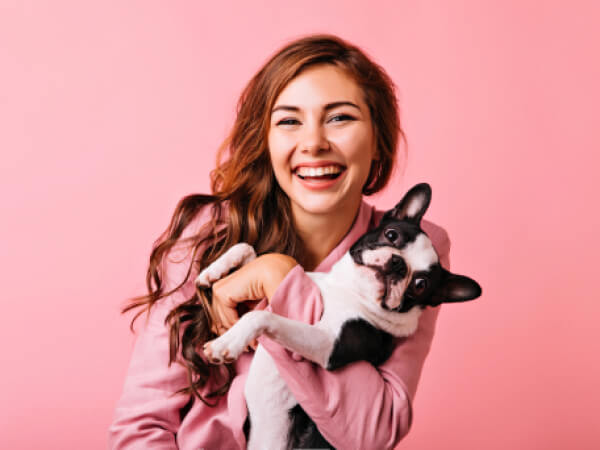 Smiling woman in pink jacket holding a black and white French bulldog against a pink background.