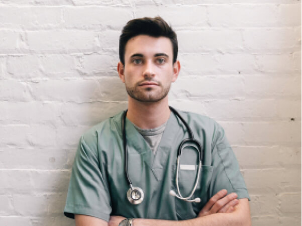 Young male healthcare professional in green scrubs with stethoscope against a white brick wall.