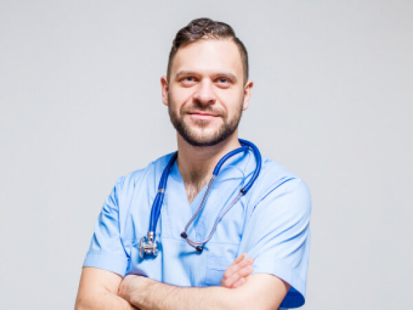 Male doctor with stethoscope around neck wearing light blue scrubs, arms crossed, smiling at camera.