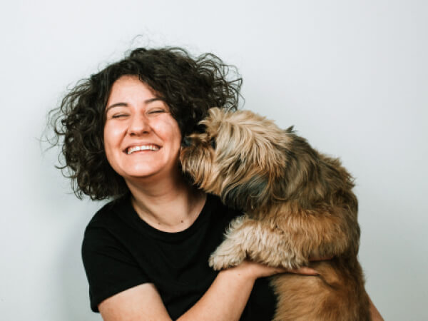 Smiling woman with curly hair holding a small fluffy dog licking her cheek against a light background.