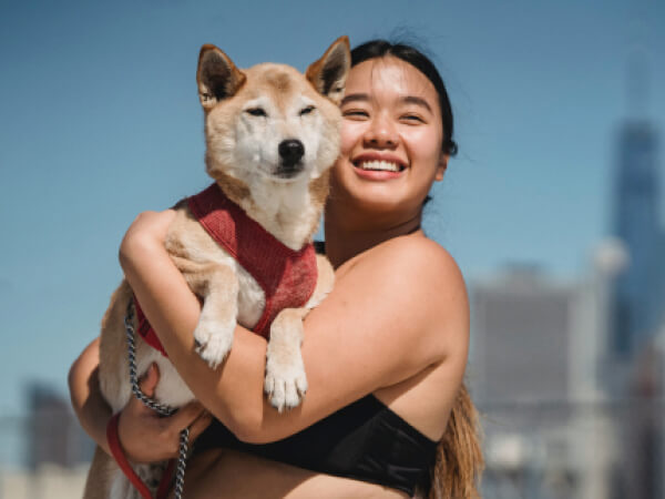 Smiling woman holding a Shiba Inu dog wearing a red harness against a city skyline background.