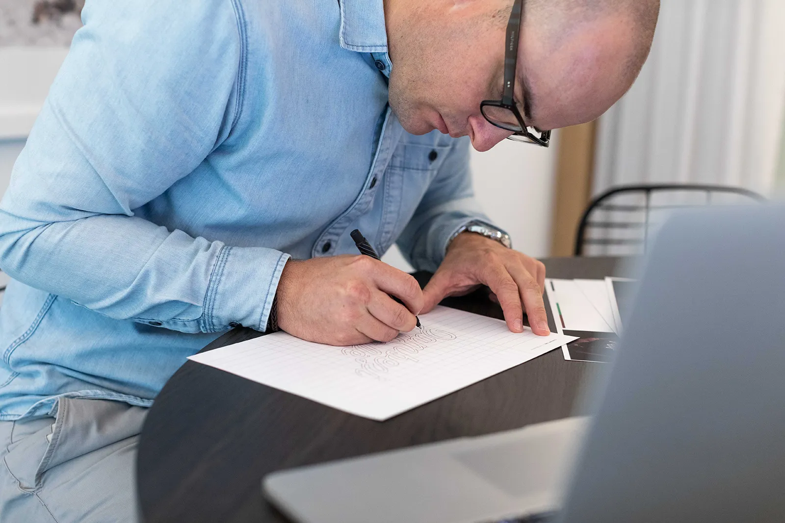 Rory in a light blue shirt sits at a table, carefully sketching lettering on lined paper with a black pen. A laptop and some printed materials are nearby, suggesting a creative design or branding process.