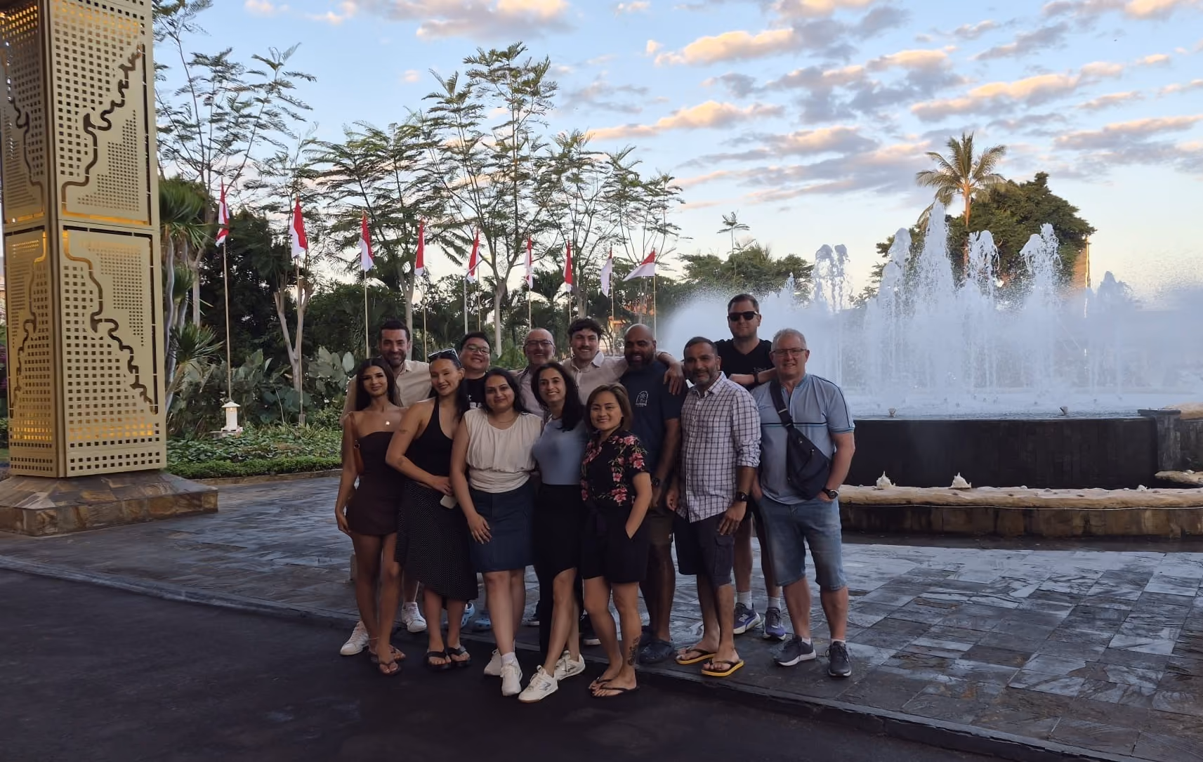 Group of diverse people posing together on a wooden deck near a reflective pool with a large tree decorated with string lights behind them during sunset.