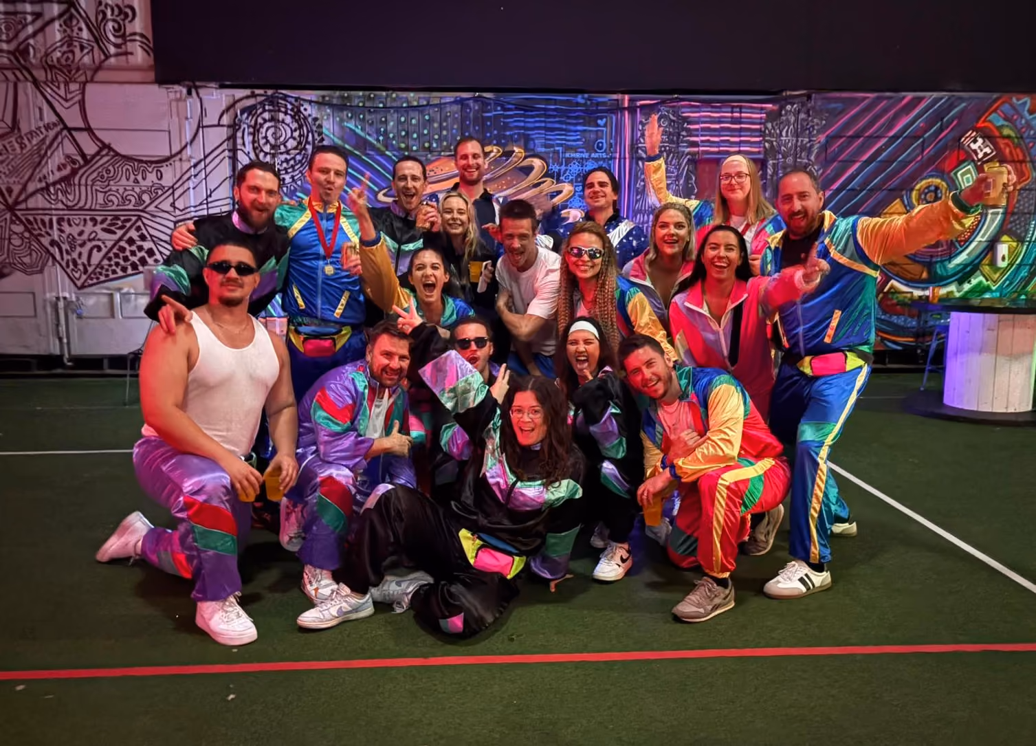 Group of 18 diverse coworkers smiling and posing together in a modern office space with glass walls and a dartboard on the wall.