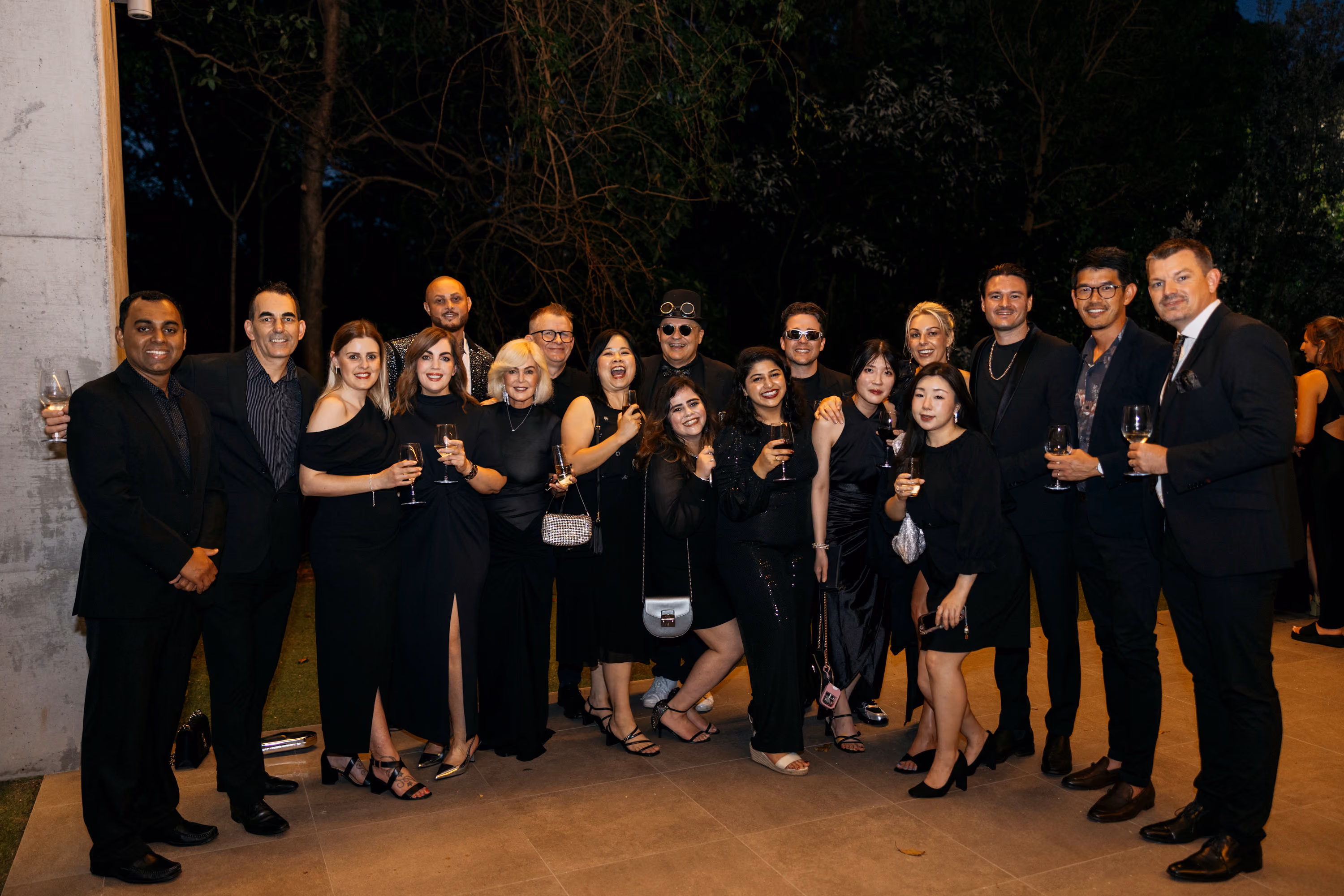 Group of nine smiling people wearing black Talent brand shirts and sunglasses outdoors on a sunny day.