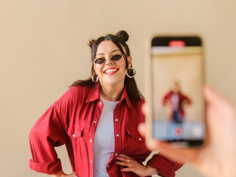Girl with sunglasses on and red shirt standing in front of an iPhone filming her on TikTok