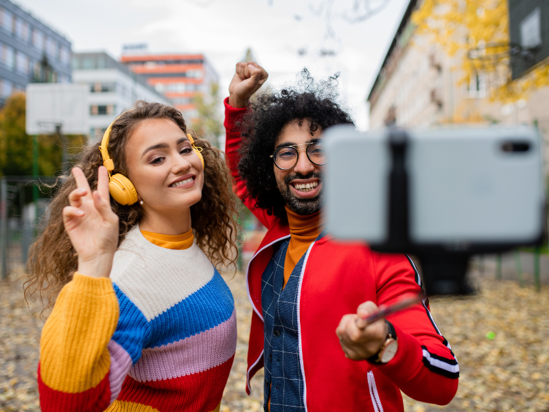 Two young people with curly hair and bright seaters dancing in front of a selfie stick with an iphone creating content for social media