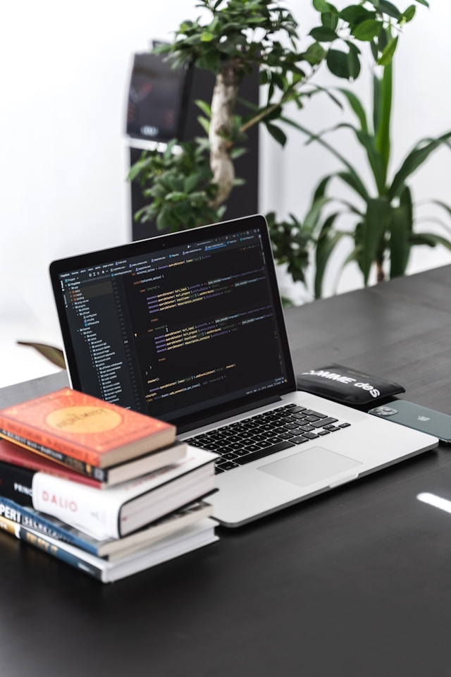 Image of a laptop with programming code on the screen, resting on a dark surface with several books stacked underneath and a plant in the background.
