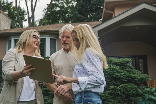 A real estate agent showing a clipboard to a couple standing outside a house with greenery around.