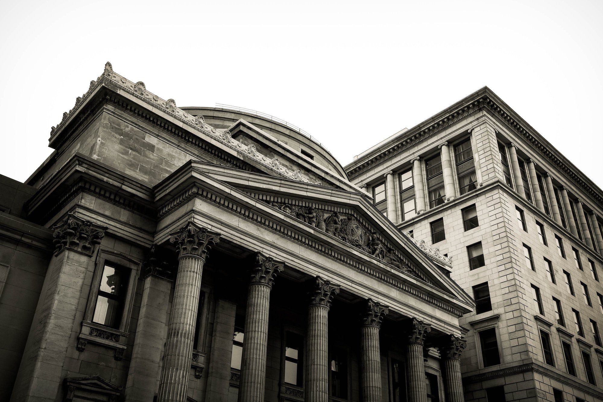 Low-angle view of a classical building with Corinthian columns and ornate pediment in front of a tall office building.
