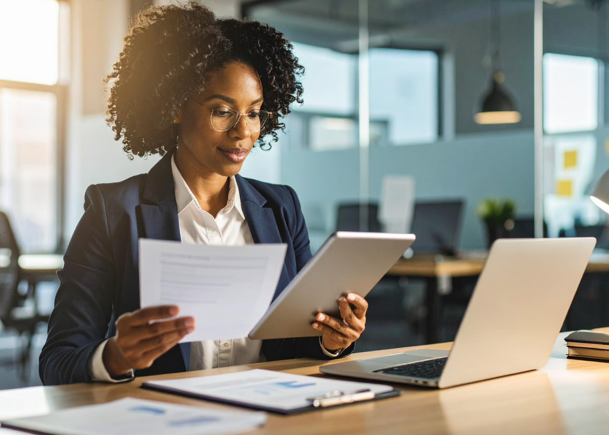 Businesswoman in glasses reviewing document and tablet at office desk with laptop.