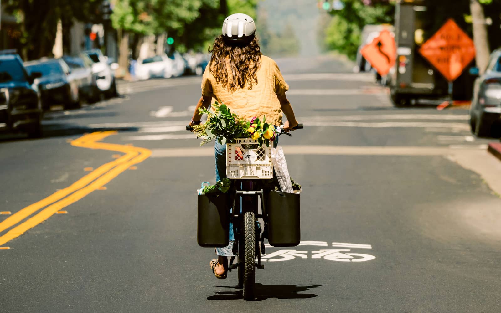 Rider wearing an ALSO Alpha Wave helmet on an ALSO TM-B e-bike in a city bike lane, carrying flowers and groceries in a front crate and rear cargo buckets.