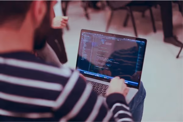 A person in a striped shirt is coding on a laptop with visible lines of code on the screen, seated in a chair in a room with others present in the background.