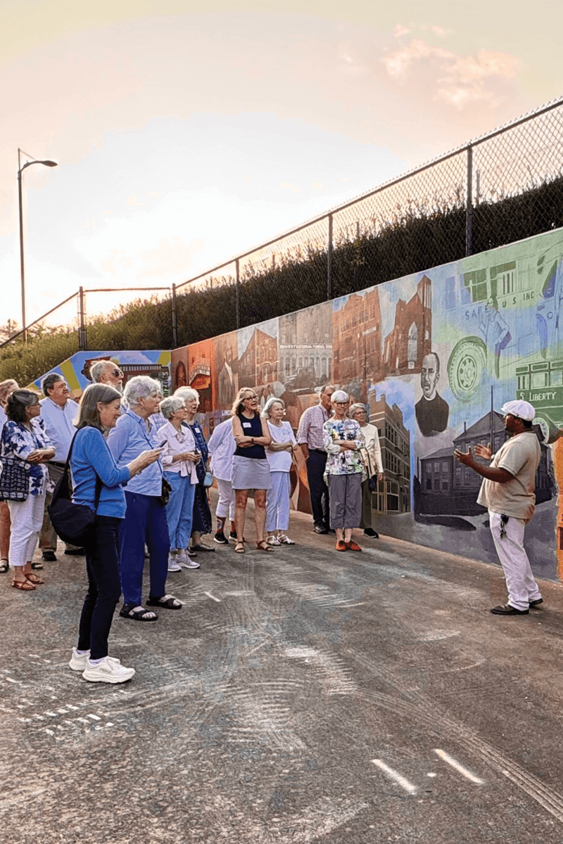 Group listening to a man speak in front of a mural