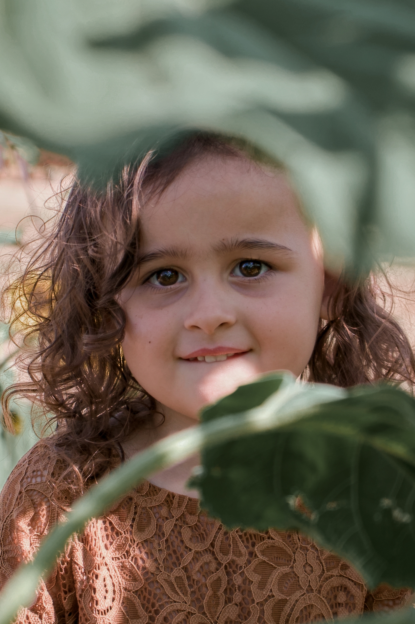 A cute little girl peeking out behind a large sunflower stock.