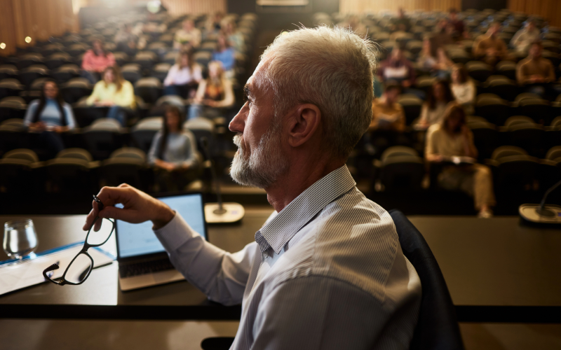 Person seated at a conference table holding glasses while observing an audience, representing attention, decision‑making, and cognitive processing.