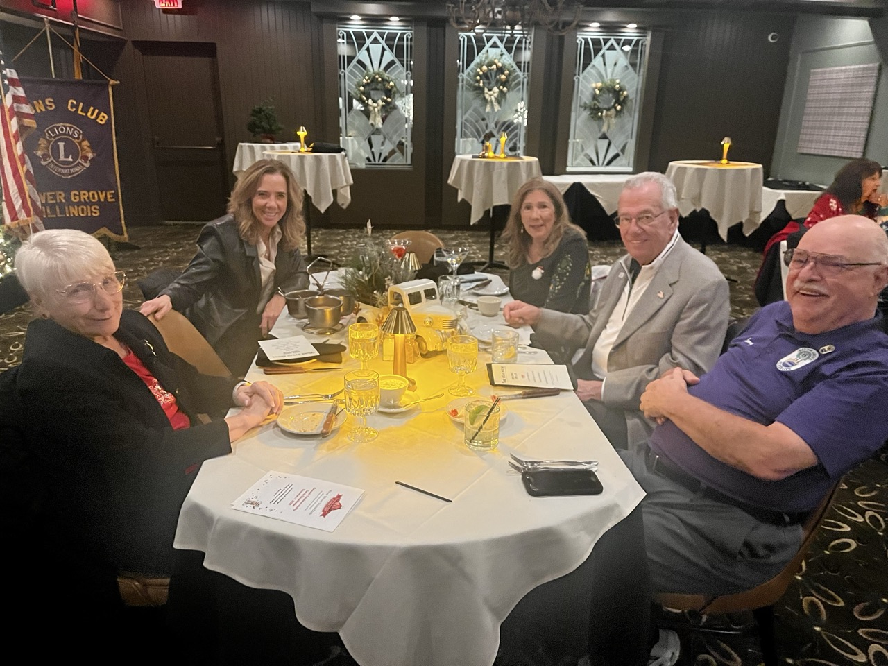 Group of smiling attendees enjoying dinner at a round table during the River Grove Lions Club 2025 holiday dinner, with festive decorations, chandeliers, wreaths, and a Lions Club banner in the background; tables set with white linens, candles, appetizers, drinks, and menus. (River Grove Lions Club holiday event)