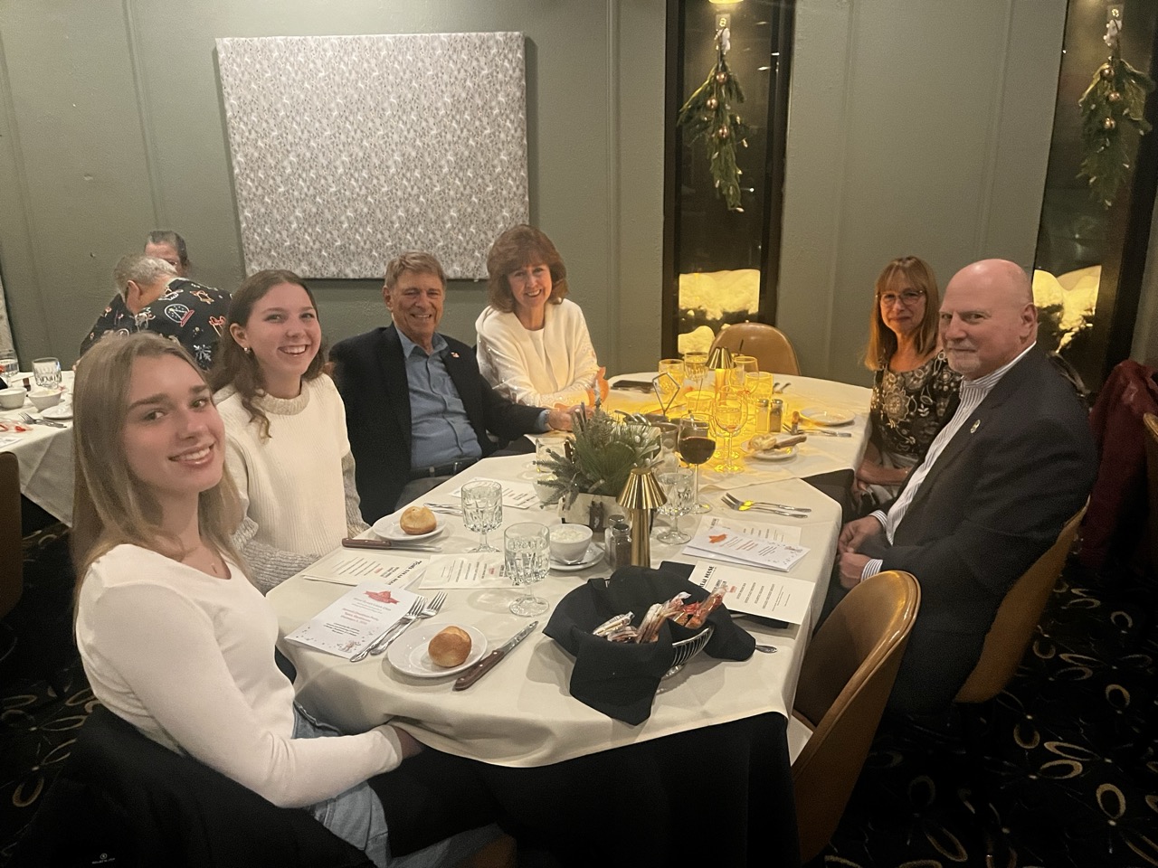 Group of smiling attendees seated at a round table during the River Grove Lions Club 2025 holiday dinner, enjoying appetizers, bread rolls, drinks, and festive centerpieces with greenery and gold accents; mistletoe and decorative wall art in the background of the warmly lit banquet room. (River Grove Lions Club holiday event)
