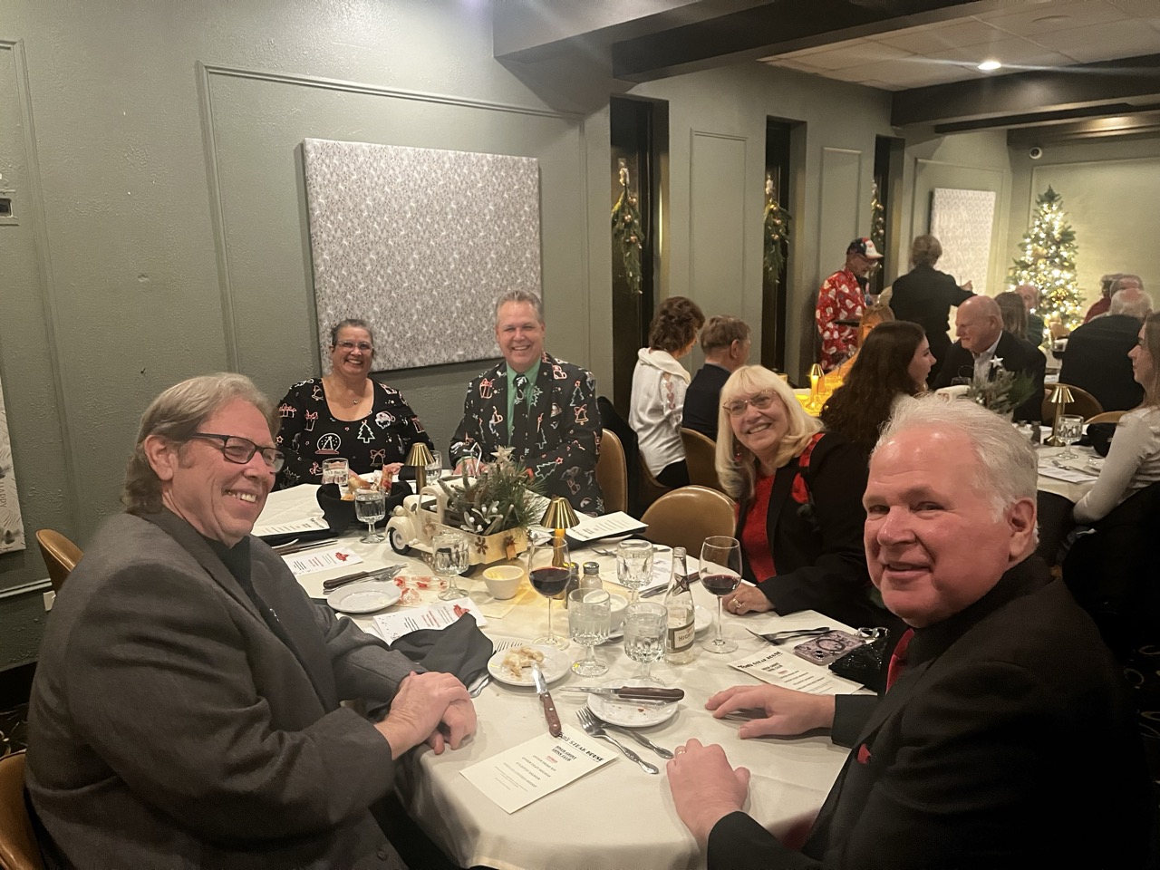 Group of smiling attendees seated at a round table during the River Grove Lions Club 2025 holiday dinner, dressed in festive attire with holiday sweaters and suits; table set with appetizers, wine, centerpieces featuring a toy car and greenery, menus, and glassware in a banquet room with Christmas tree and wall art in the background. (River Grove Lions Club holiday event)