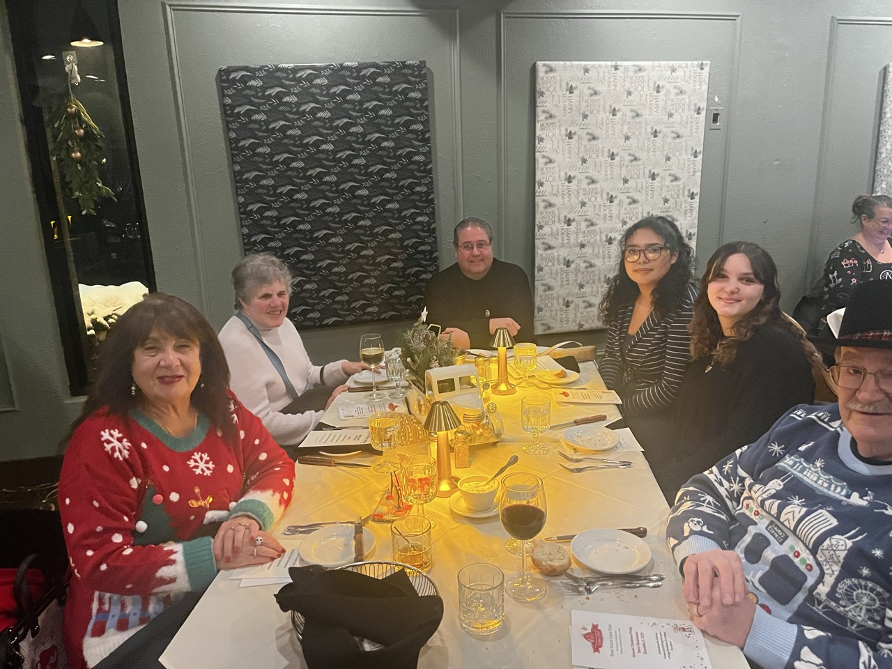 Group of smiling attendees in festive holiday sweaters seated around a table during the River Grove Lions Club 2025 holiday dinner, enjoying wine, appetizers, bread, and glowing centerpieces with gold lamps and greenery; mistletoe and patterned wall art in the background of the banquet room. (River Grove Lions Club holiday event)