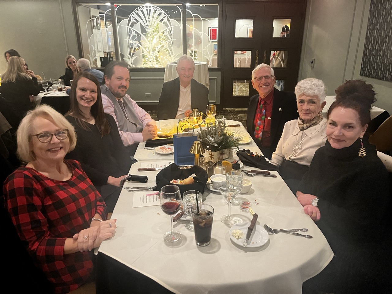Group of smiling attendees seated at a long table during the River Grove Lions Club 2025 holiday dinner, in festive and formal attire with holiday sweaters, suits, and pins; table set with wine, appetizers, bread, glowing centerpieces, and menus in a banquet room featuring ornate stained-glass style window decorations and wall art. (River Grove Lions Club holiday event)