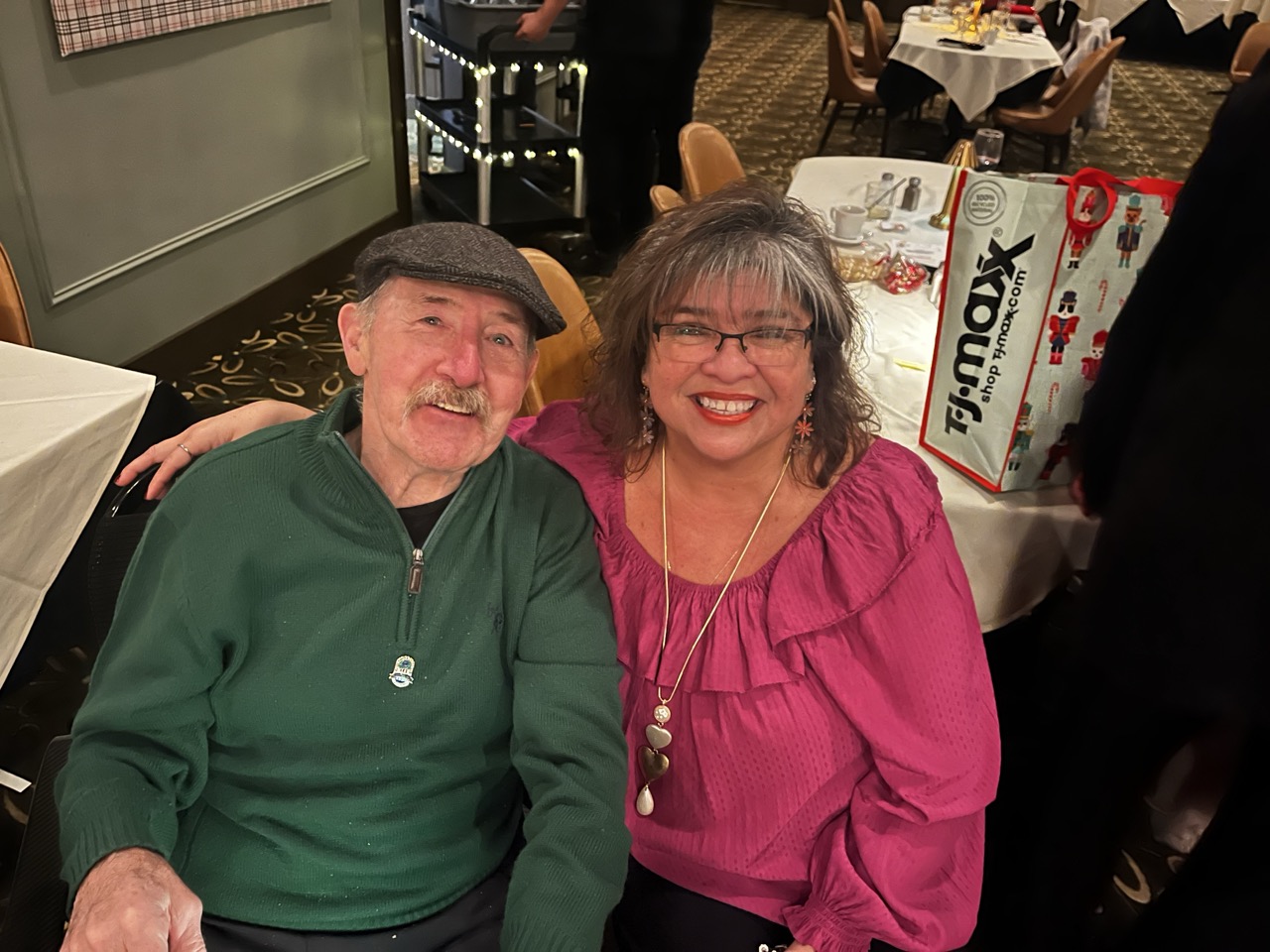 Smiling man in green jacket and flat cap seated next to a woman in a pink ruffled top during the River Grove Lions Club 2025 holiday dinner; her arm around his shoulder, with festive gift bag, table settings, and twinkling lights on serving carts in the banquet room background. (River Grove Lions Club holiday event)
