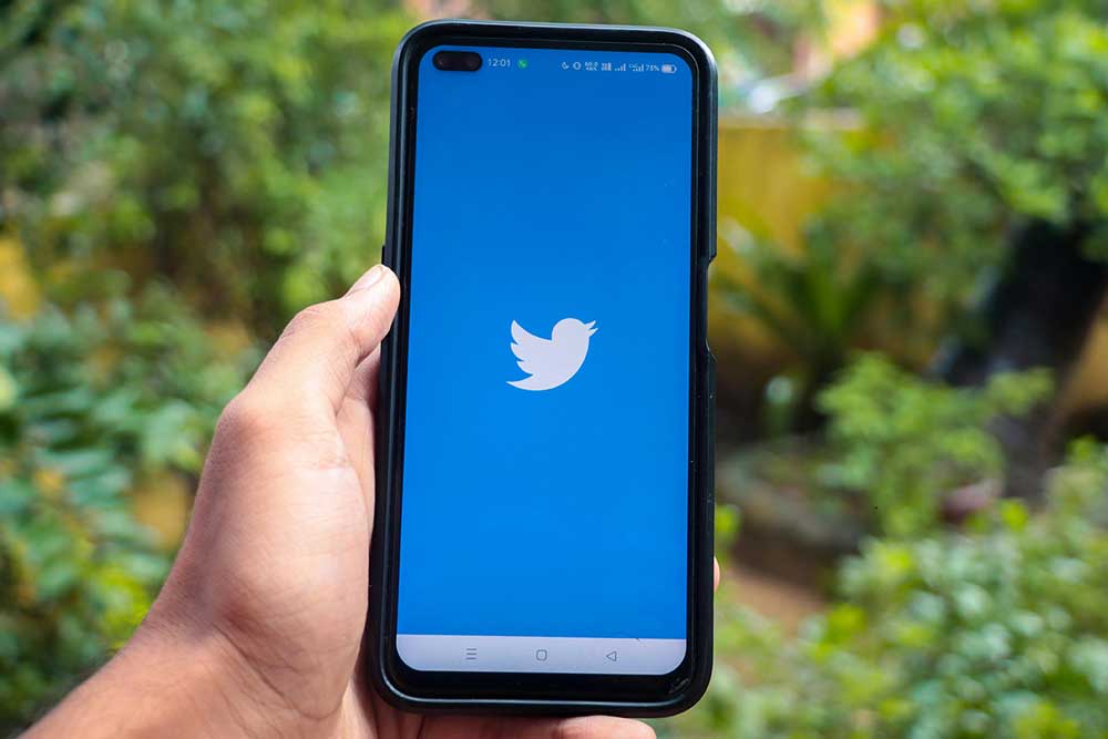 Hand holding a mobile phone. The screen of the phone shows the Twitter bird icon against a blue background.