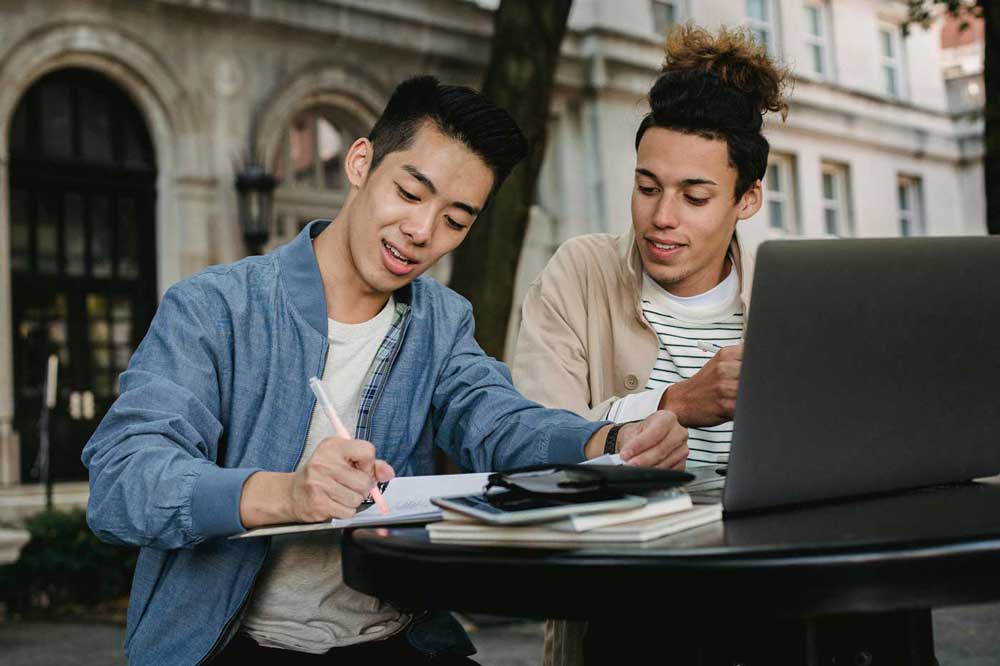 Two young men work together on a laptop at a table outside.