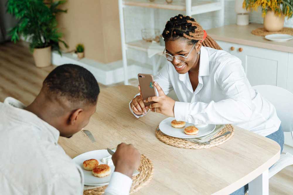Woman uses her phone to photograph a man eating across the table from her.