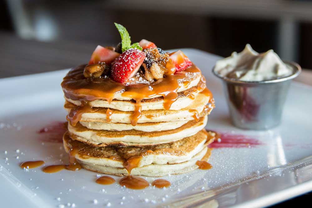 A stack of pancakes covered in gooey syrup, powdered sugar, and fresh fruit on a white ceramic plate.