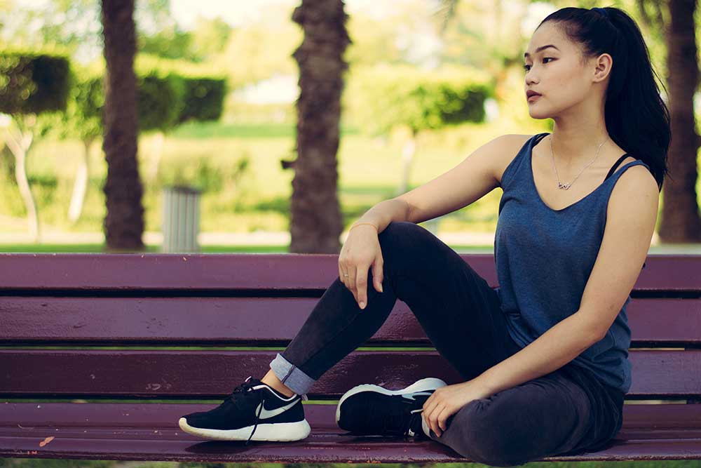 A young woman sits on a purple park bench. She wears lightweight athleisure clothing and black Nike running shoes.