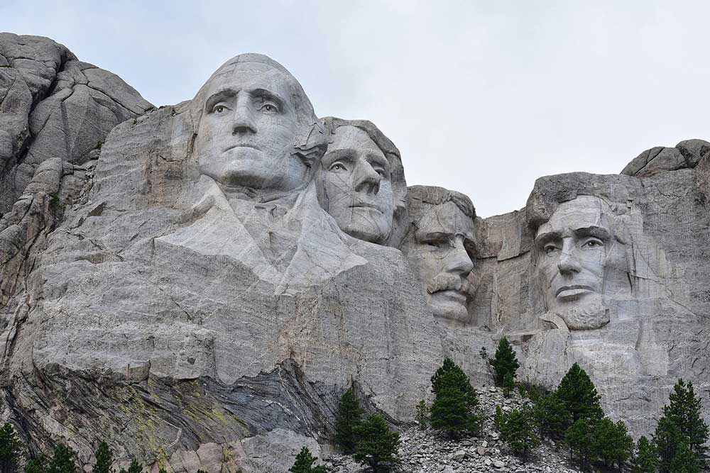 Mount Rushmore is shown under a hazy blue sky. The cliffside in front of it is dotted with evergreen trees.
