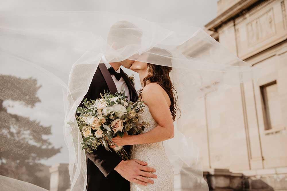 A newlywed couple kisses. The groom wears a black tuxedo, and the bride wears a white wedding dress and veil that swirls around the couple as they embrace. She also holds an elaborate bouquet of flowers.