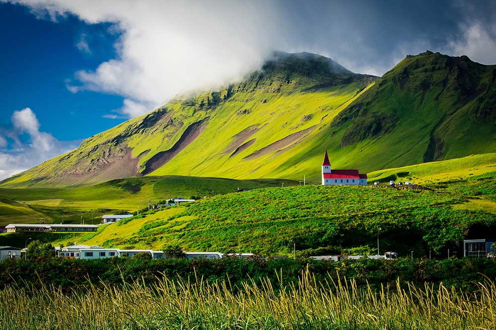 The Vík i Myrdal Church, a small white structure with a red roof and a single steeple, sits on a green hilltop in the distance overlooking a small town on a sunny day in Iceland. Beyond the church are idyllic green mountain peaks surrounded by fog.